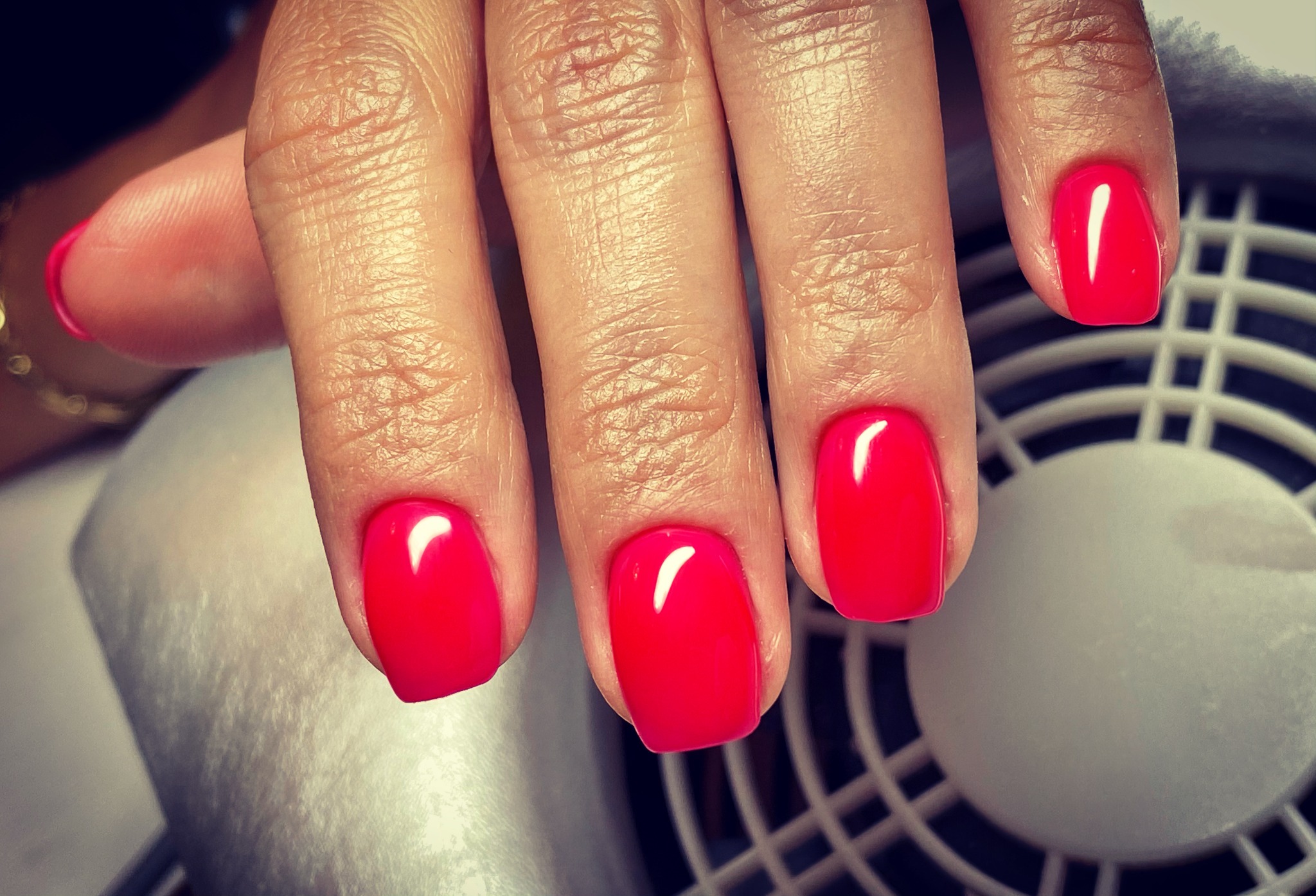 Dłoń z jaskrawoczerwonym, kwadratowym manicure hybrydowym na tle pochłaniacza pyłu.