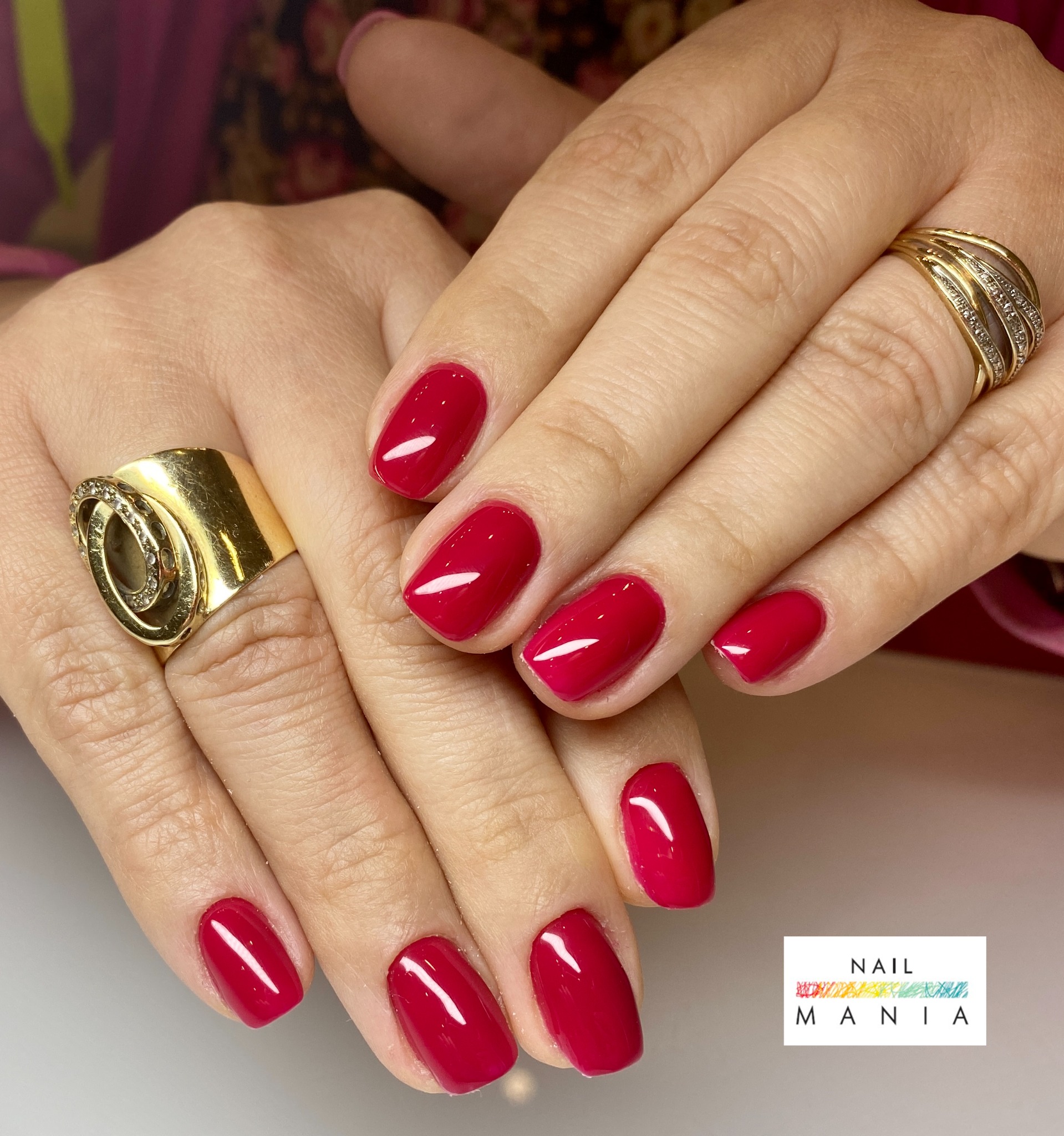 Dłonie kobiety z czerwonym, błyszczącym manicure hybrydowym i złotymi pierścionkami, prezentacja efektu zabiegu.