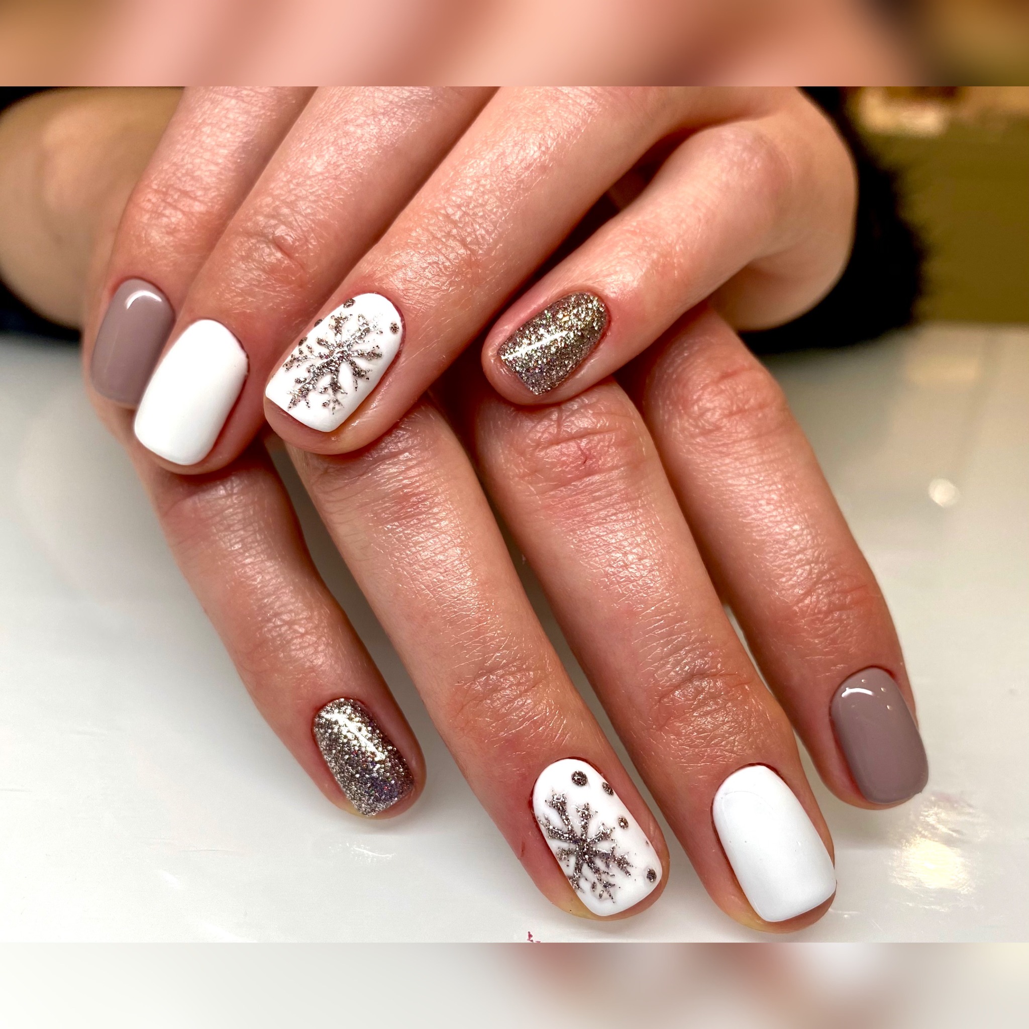 Dłonie z zimowym manicure: paznokcie w kolorach bieli, taupe i brokatu, zdobione srebrzystymi płatkami śniegu.
