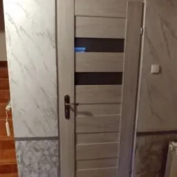 Wykończone wnętrze: drzwi wewnętrzne z przeszkleniami na tle ściany z efektem marmuru, z dekoracyjnym pasem imitującym beton u dołu. Widoczna część schodów i grzejnik.