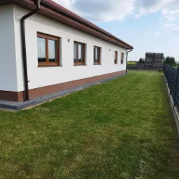 Ocieplony budynek jednorodzinny z brązowymi oknami i dachem, widok z boku na zadbany trawnik i ogrodzenie panelowe.