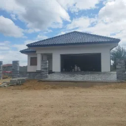 Nowoczesny dom jednorodzinny z białymi ścianami i grafitowym dachem, wybrukowany podjazd z kostki, w tle błękitne niebo z chmurami, widoczne elementy ogrodzenia z szarej kostki, teren wokół domu...