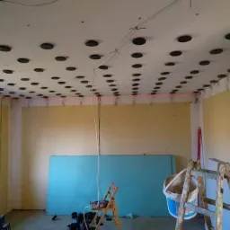 Przygotowanie sufitu podwieszanego: widoczne profile montażowe, płyta karton-gips oparta o ścianę, poziomica laserowa i drabina w pomieszczeniu z żółtymi ścianami.