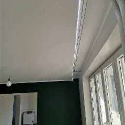 Montaż listwy oświetleniowej LED przy suficie podwieszanym, widok na okno z białymi ramami i fragment ściany pomalowanej na ciemny zielony kolor.