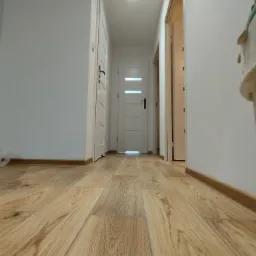 Perspektywa z poziomu podłogi na korytarz z ułożonymi panelami podłogowymi w odcieniach jasnego drewna, białe ściany i drzwi widoczne w głębi.