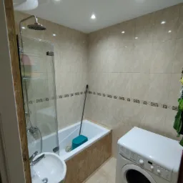 Wykończona łazienka w beżowych płytkach z kabiną prysznicową, umywalką i pralką Indesit, widoczna mop w wannie.