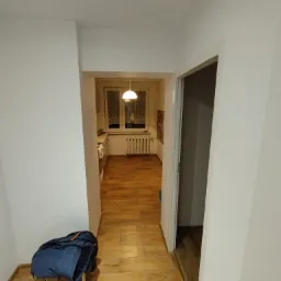 Widok na odnowione wnętrze mieszkania z jasnymi ścianami i drewnianą podłogą, z kurtką leżącą na krześle w rogu