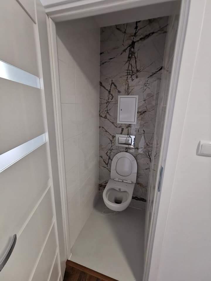 Wąska toaleta po remoncie, widok z zewnątrz, jasne płytki imitujące marmur na ścianie za muszlą, biała armatura, drzwi z białymi panelami po lewej stronie.