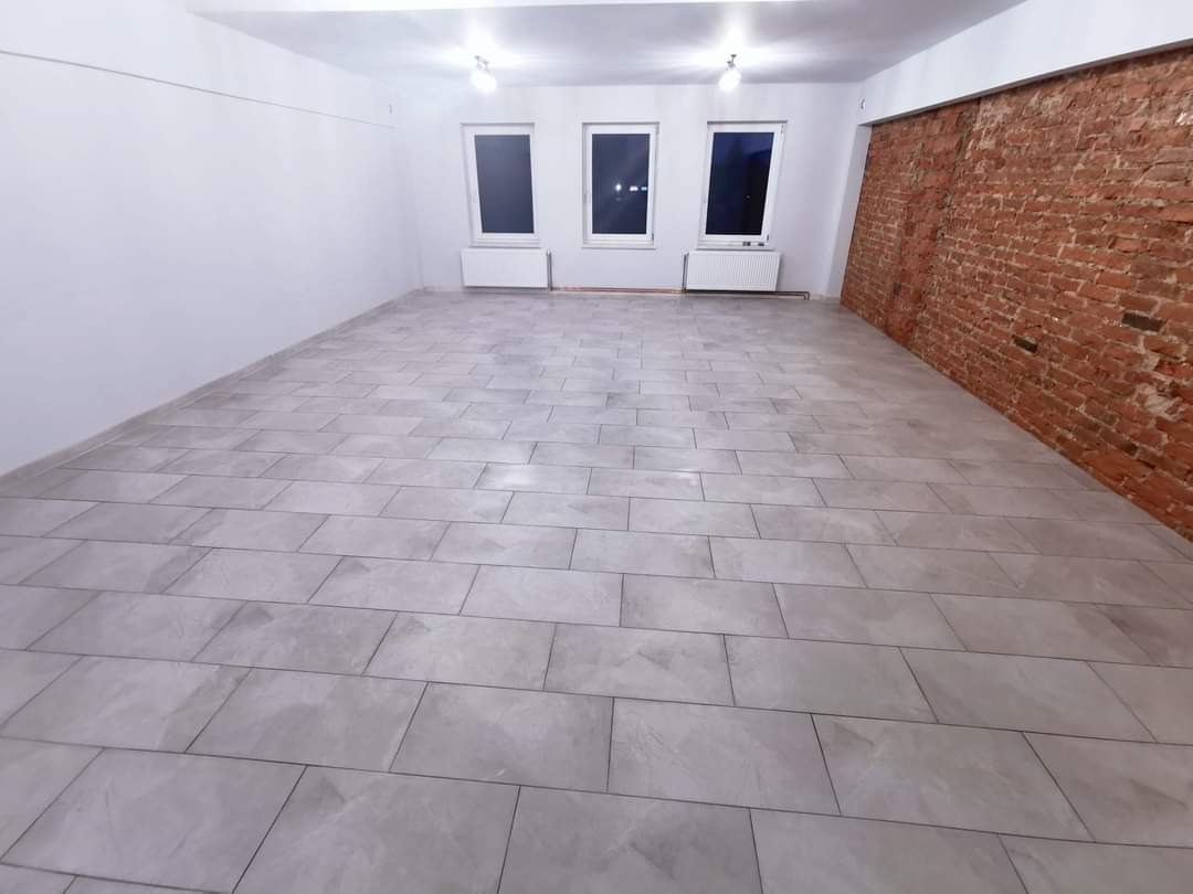 Wykończone wnętrze z szarymi płytkami podłogowymi ułożonymi w prostokątnym wzorze, białe ściany kontrastują z fragmentem ściany z czerwonej cegły, trzy okna z białymi ramami i grzejnikami pod spodem.