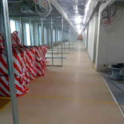 Długi korytarz w trakcie budowy z widocznymi metalowymi stelażami, pakietami płyt gipsowo-kartonowych w czerwono-białe pasy i instalacjami pod sufitem.