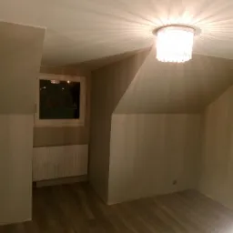 Poddasze po remoncie, świeżo pomalowane na szaro, z widocznym oknem, grzejnikiem, drewnianą podłogą i ozdobnym oświetleniem sufitowym.