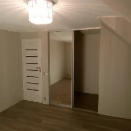Pokój na poddaszu po remoncie z drewnianą podłogą, beżowymi ścianami i szafą wnękową z lustrzanymi drzwiami przesuwnymi, oświetlony ozdobnym żyrandolem.