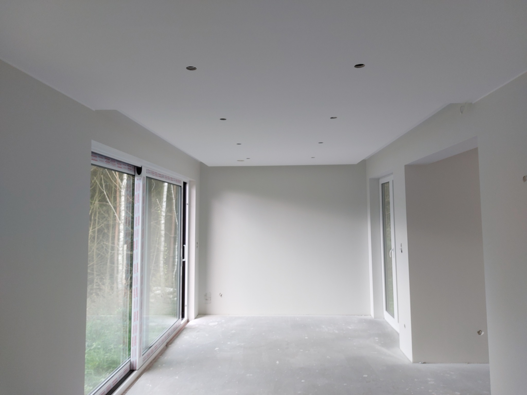 Wnętrze świeżo wykończonego pokoju z białymi ścianami, sufit z punktowymi lampami i duże przesuwne okno z widokiem na drzewa.