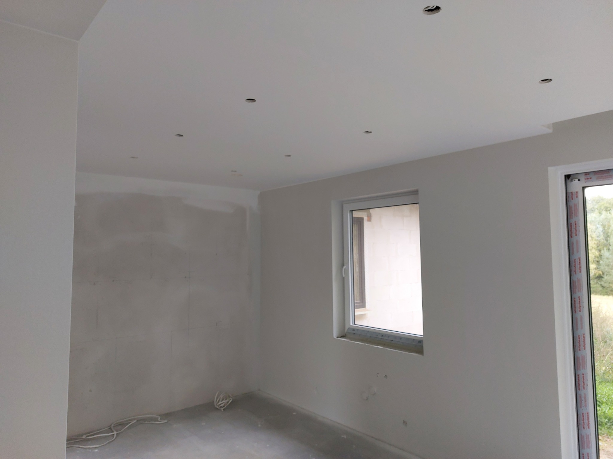 Pokój w trakcie remontu: ściany po szpachlowaniu, przygotowane do malowania, widoczne otwory pod oświetlenie w suficie, okno z widokiem na zewnątrz.