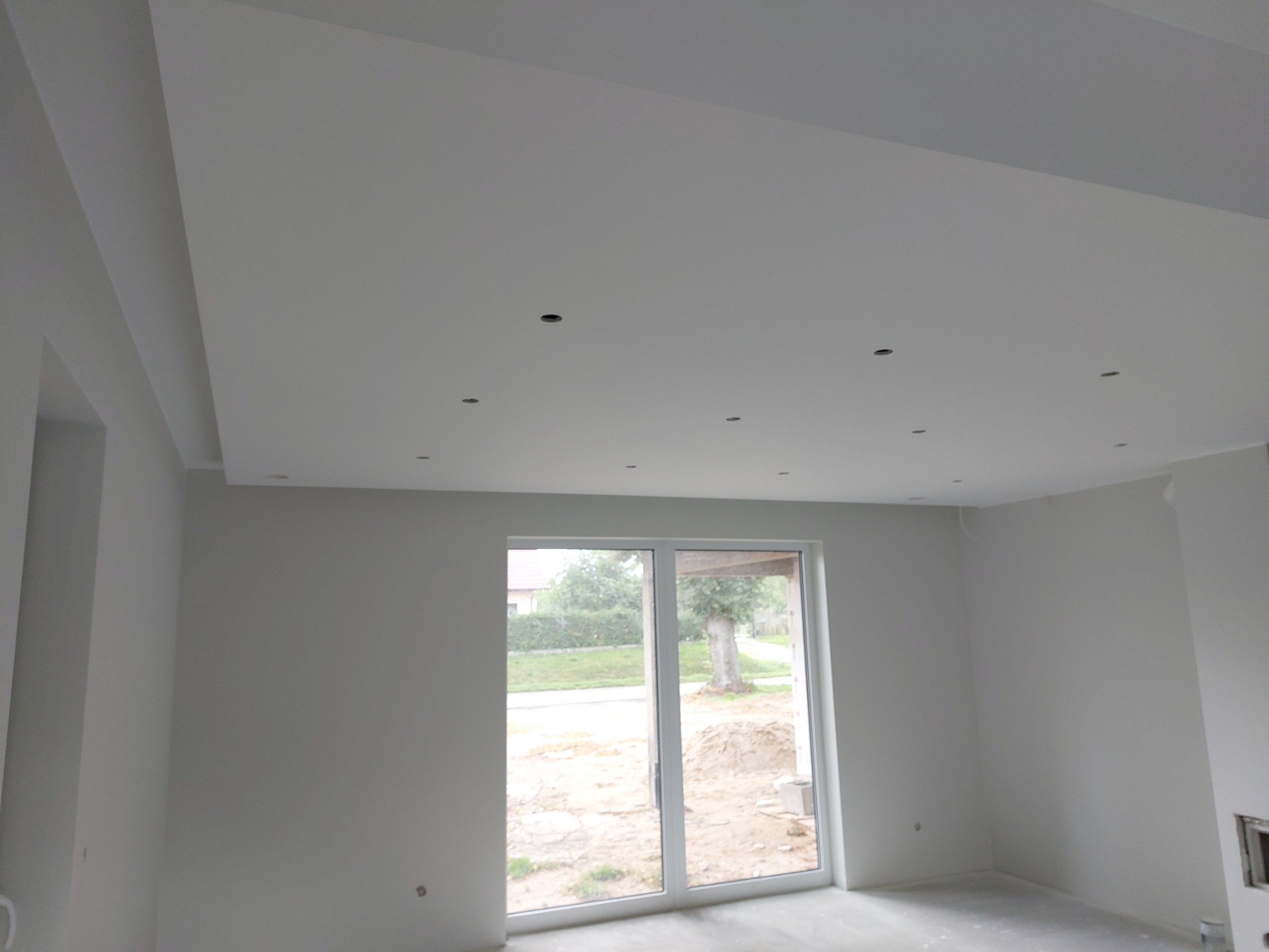 Wnętrze pokoju po remoncie z gładkimi, pomalowanymi na biało ścianami i sufitem podwieszanym z otworami na oświetlenie punktowe, widok na zewnątrz przez duże okno balkonowe.