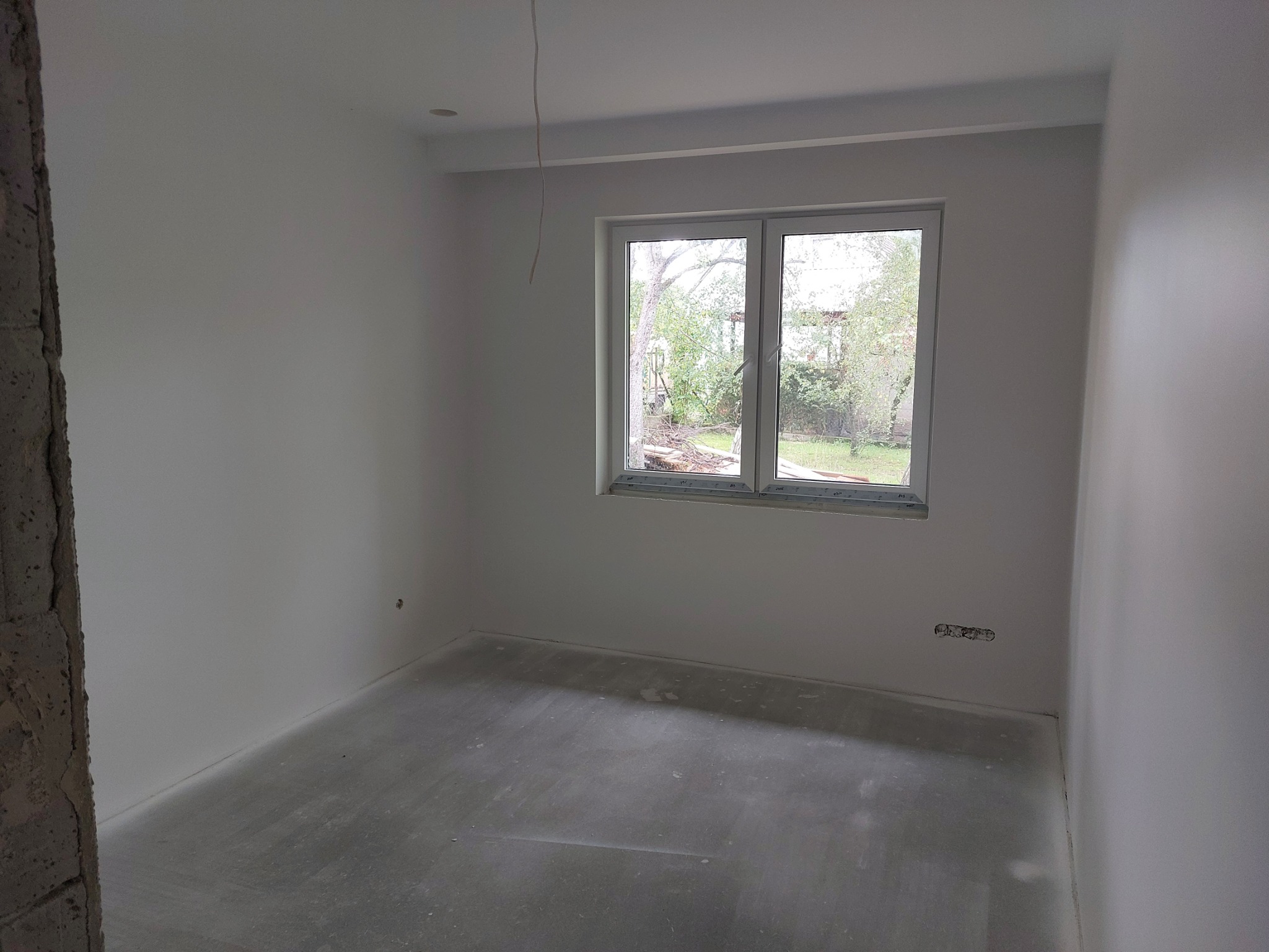 Pusty, świeżo wykończony pokój z gładkimi, białymi ścianami, szarym betonowym podłożem i białym oknem z widokiem na zieleń. Odsłonięty fragment surowego muru po lewej stronie kadru. Widoczne...