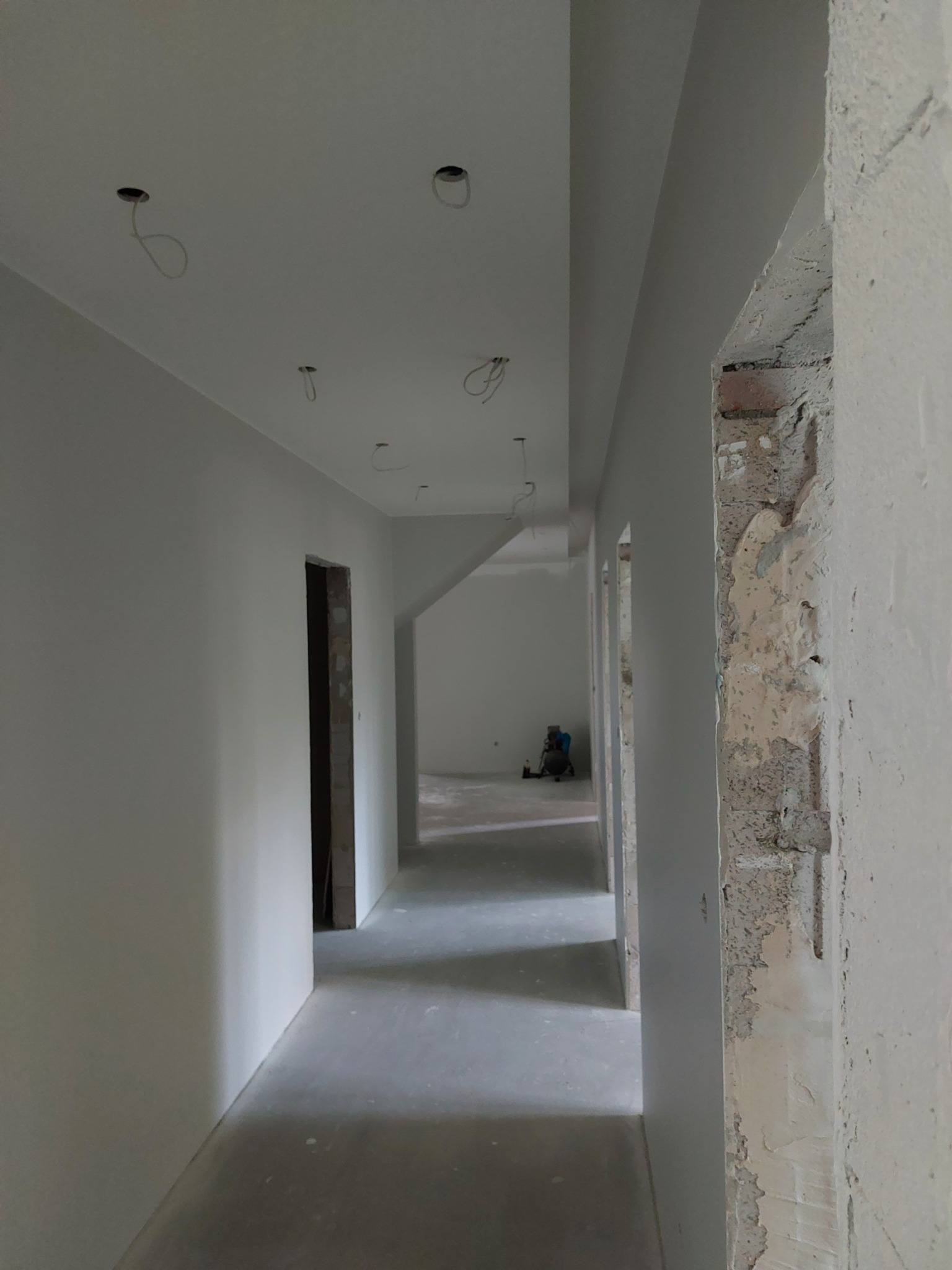 Korytarz w trakcie prac wykończeniowych, świeżo pomalowane ściany na biało, widoczne wystające kable z sufitu, surowy mur przy przejściu.