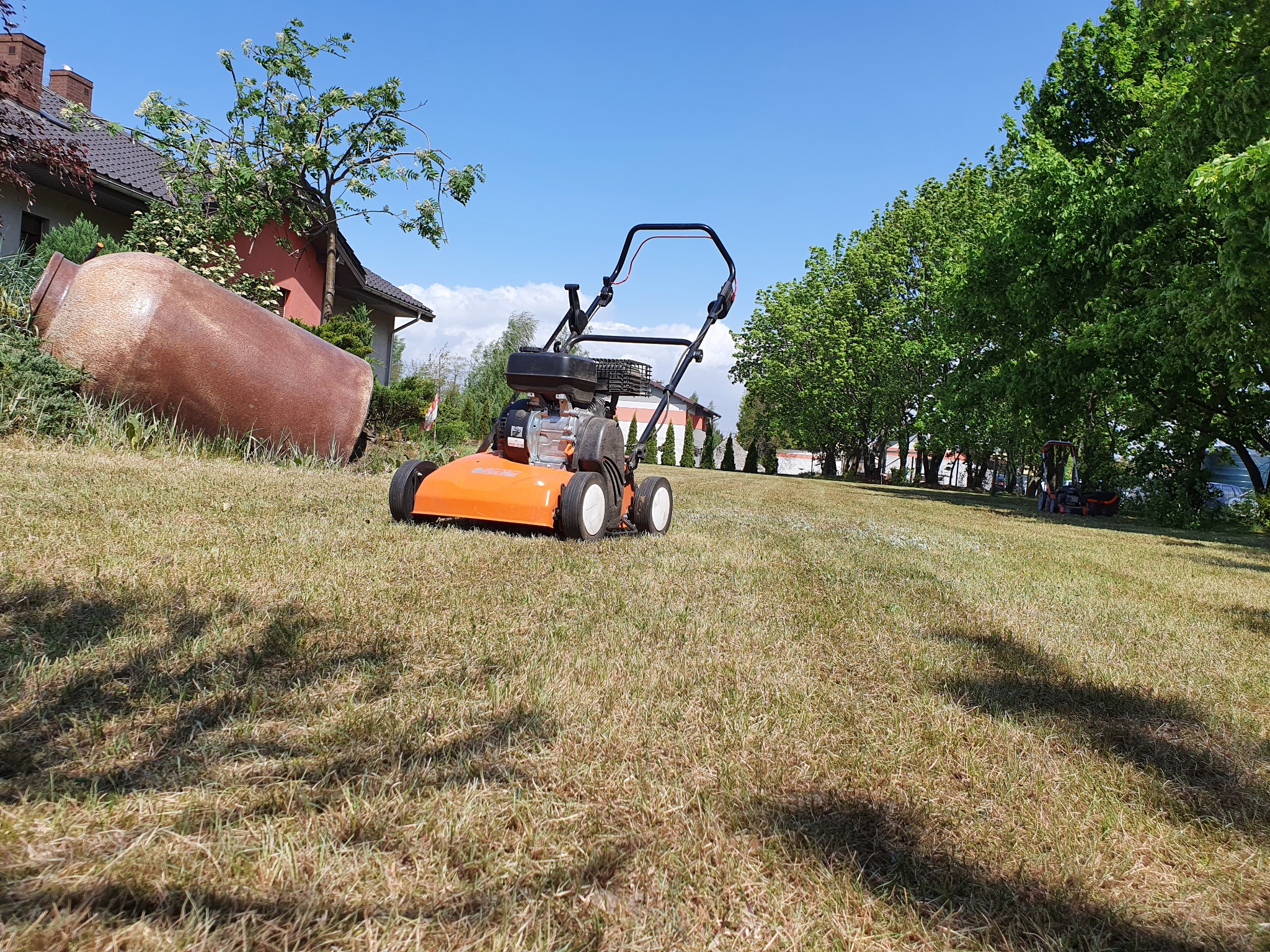 Pomarańczowa kosiarka spalinowa na trawniku z widocznymi śladami po koszeniu, w tle dom z dużą, dekoracyjną amforą i rząd drzew.