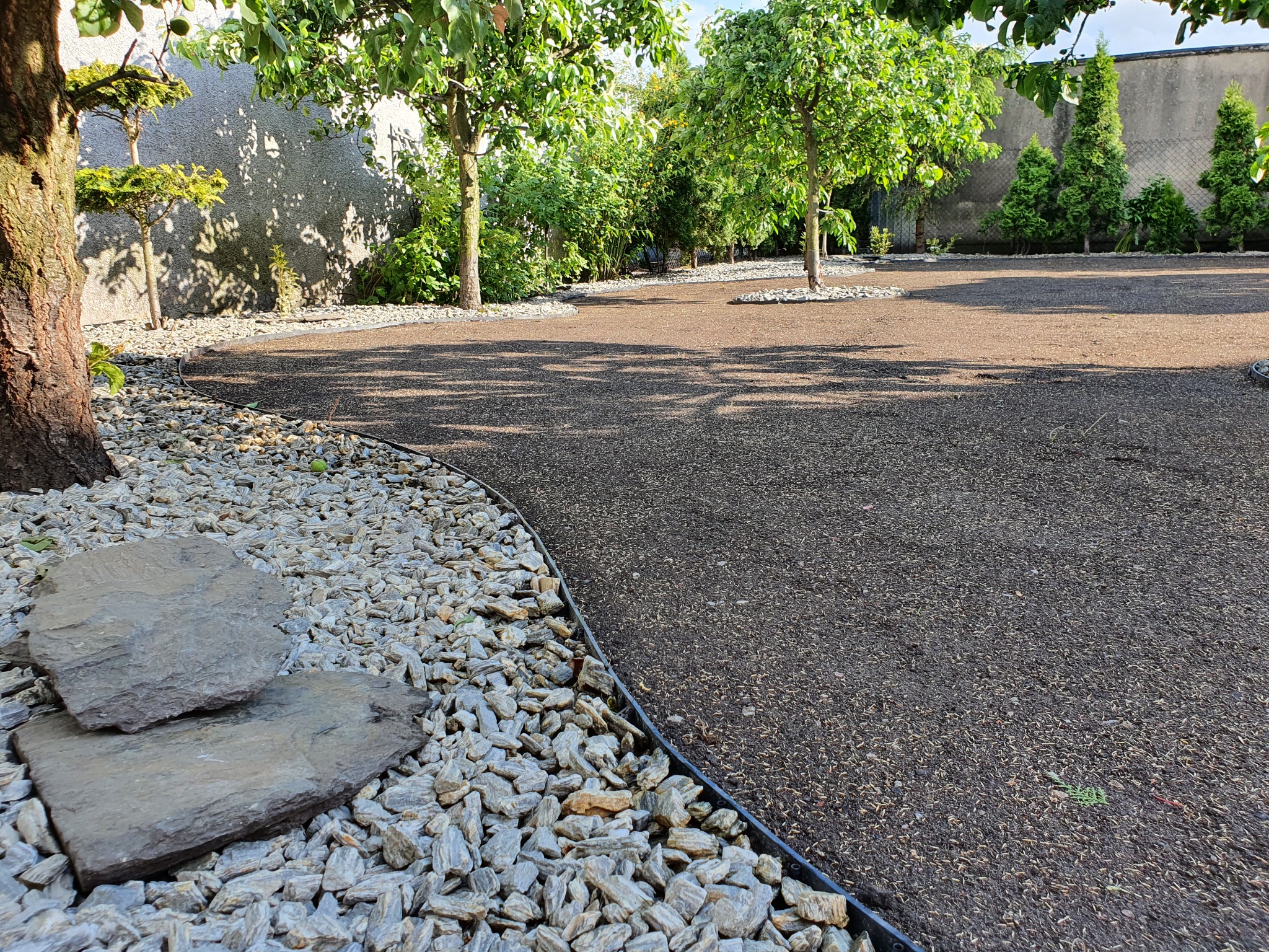 Przygotowany teren pod trawnik z widocznymi kamiennymi obrzeżami, drzewami i krzewami w tle, po wałowaniu i równaniu podłoża.