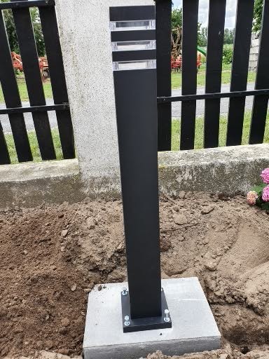 Czarna, nowoczesna lampa ogrodowa zamontowana na betonowej podstawie, otoczona ziemią i fragmentem ogrodzenia z widocznym tłem zieleni.