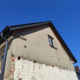 Fragment elewacji budynku z widocznymi śladami po demontażu elementów oraz nową, ciemną blachodachówką i orynnowaniem na tle błękitnego nieba.