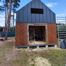 GOL MAR - Budowa małego domu z ciemnym metalowym dachem i drewnianą elewacją, widoczny otwór okienny w dachu i ekipa w środku w trakcie przerwy.