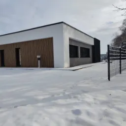 Nowoczesny dom jednorodzinny z płaskim dachem i drewnianą elewacją w zimowej scenerii. Minimalistyczny design, białe ściany, czarne akcenty, ogrodzenie panelowe.