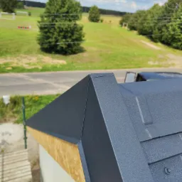 Szczegółowe ujęcie fragmentu nowo krytego dachu z blachy na budynku z widocznym deskowaniem i elementami mocującymi na tle zielonego krajobrazu.