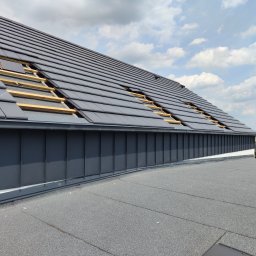 GOL MAR - Częściowo pokryty dach z ciemnej dachówki ceramicznej, widoczne drewniane łaty, błękitne niebo z obłokami w tle. Fragment pokrycia papą na płaskim dachu.