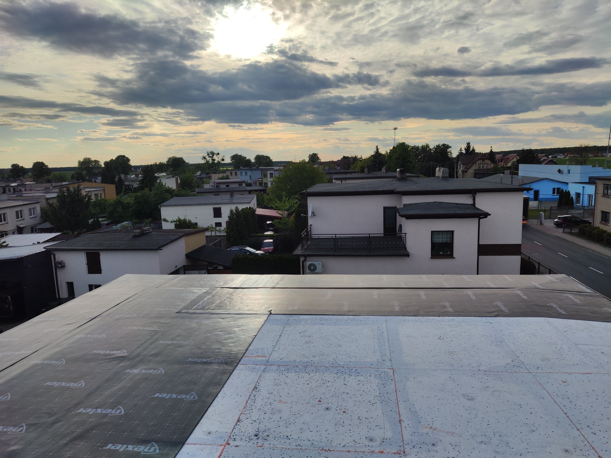 Fragment dachu w trakcie prac, widoczne warstwy izolacyjne, na tle zabudowań mieszkalnych i pochmurnego nieba oświetlonego słońcem.