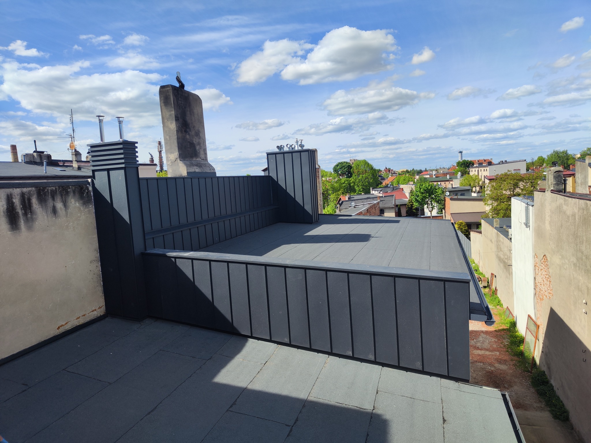 Nowoczesny, płaski dach z blachy na budynku miejskim, widok z góry na tle błękitnego nieba i zabudowy miejskiej. Ciemnoszara kolorystyka dachu.