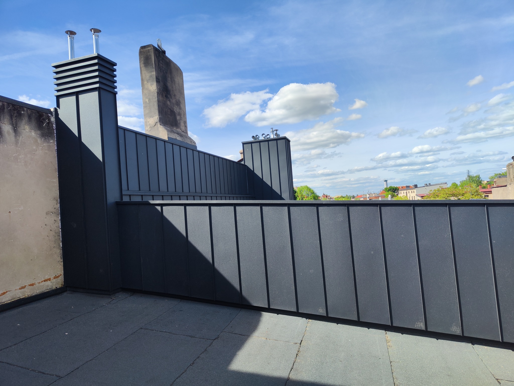 Szare, metalowe obłożenie dachu z kominami i widokiem na miasto w tle, pod błękitnym niebem z chmurami.