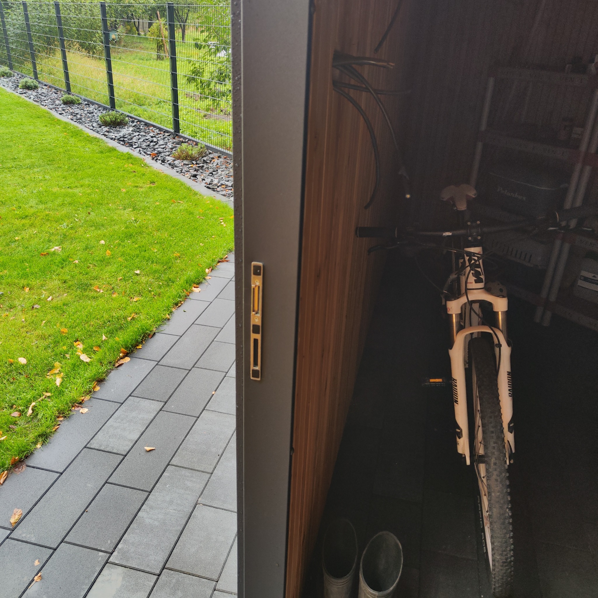 Otwarty garaż z drewnianym wykończeniem, widoczny rower górski, półki i gumiaki. Na zewnątrz trawnik i ścieżka z kostki brukowej, oddzielone metalowym ogrodzeniem.