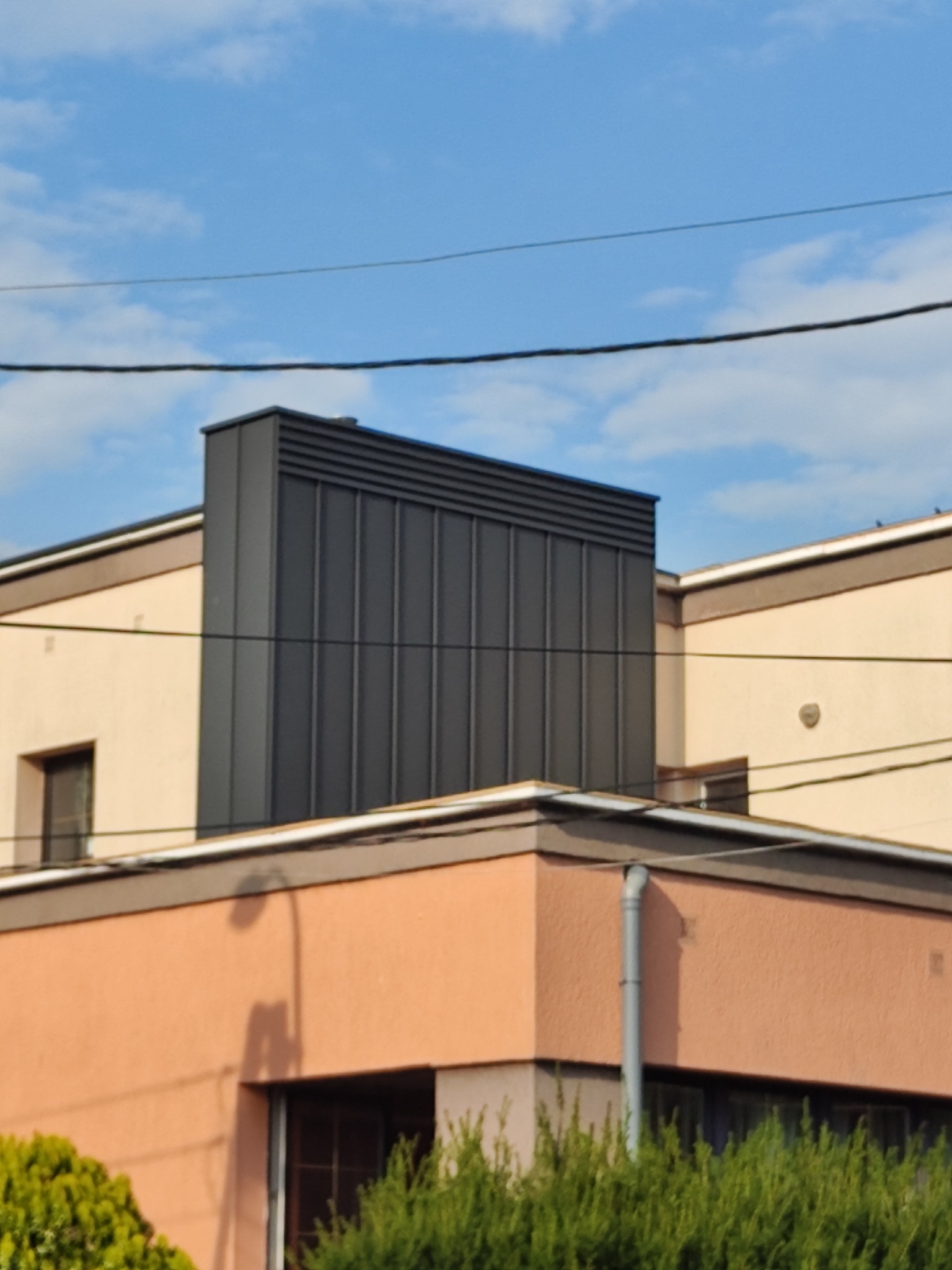 Nowoczesna obróbka blacharska komina na dachu budynku mieszkalnego, widoczna na tle błękitnego nieba z liniami energetycznymi.