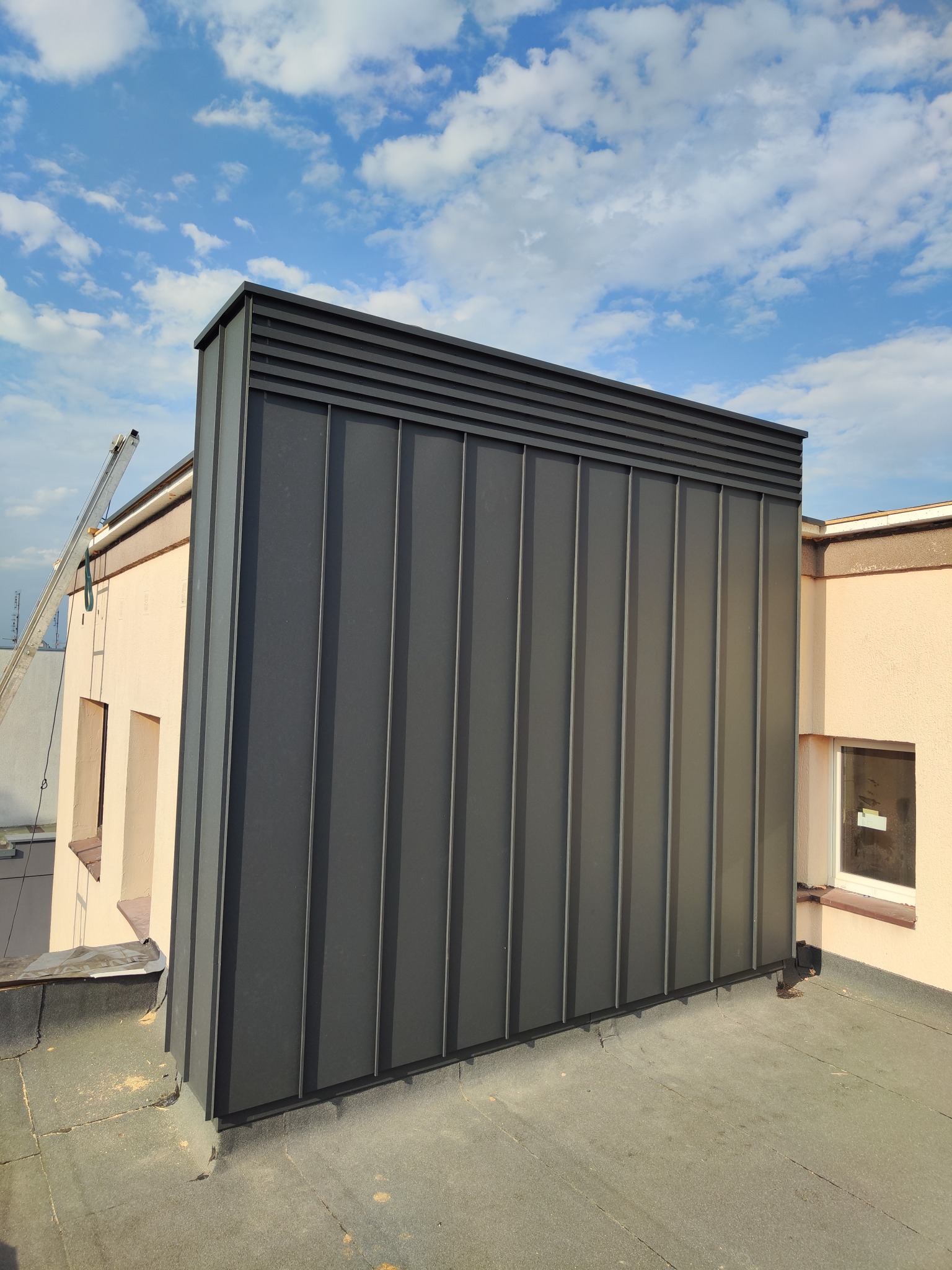 Szara obudowa komina z blachy na dachu budynku, widok z dołu. Na drugim planie fragment elewacji i drabina oparta o ścianę. Błękitne niebo z chmurami w tle.