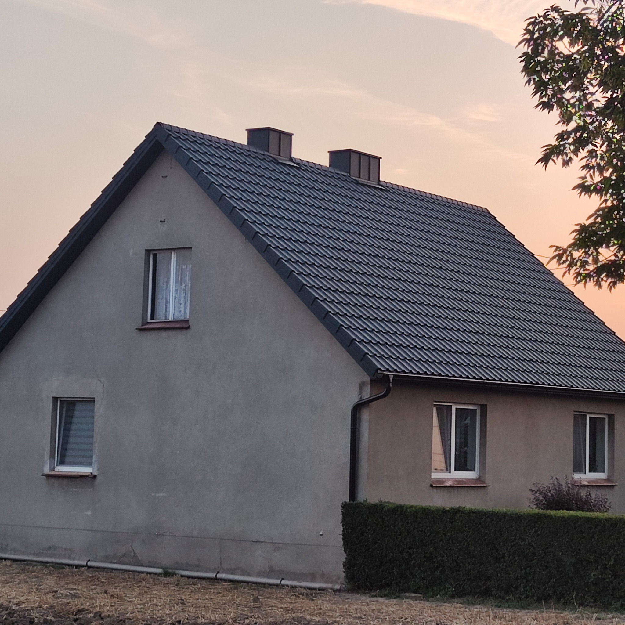 Dom jednorodzinny z nowym, ciemnym dachem w odcieniach szarości, widoczny z boku, z drzewem po prawej stronie, sfotografowany o zmierzchu przy pomarańczowym niebie.