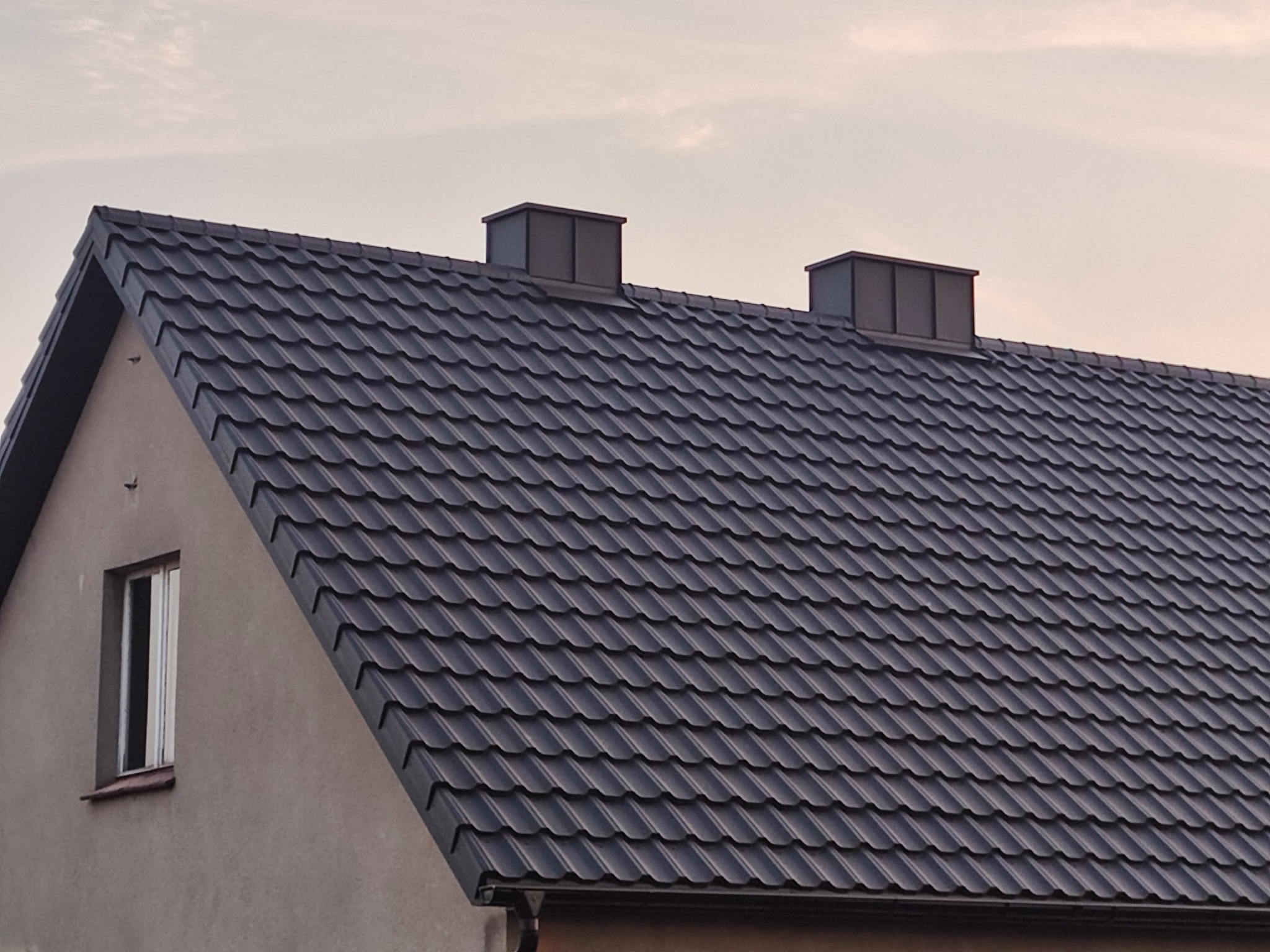 Ciemny dach z blachy trapezowej na domu jednorodzinnym, widoczne dwa kominy wentylacyjne i okno. Dach w odcieniach szarości i czerni, na tle jasnego nieba.