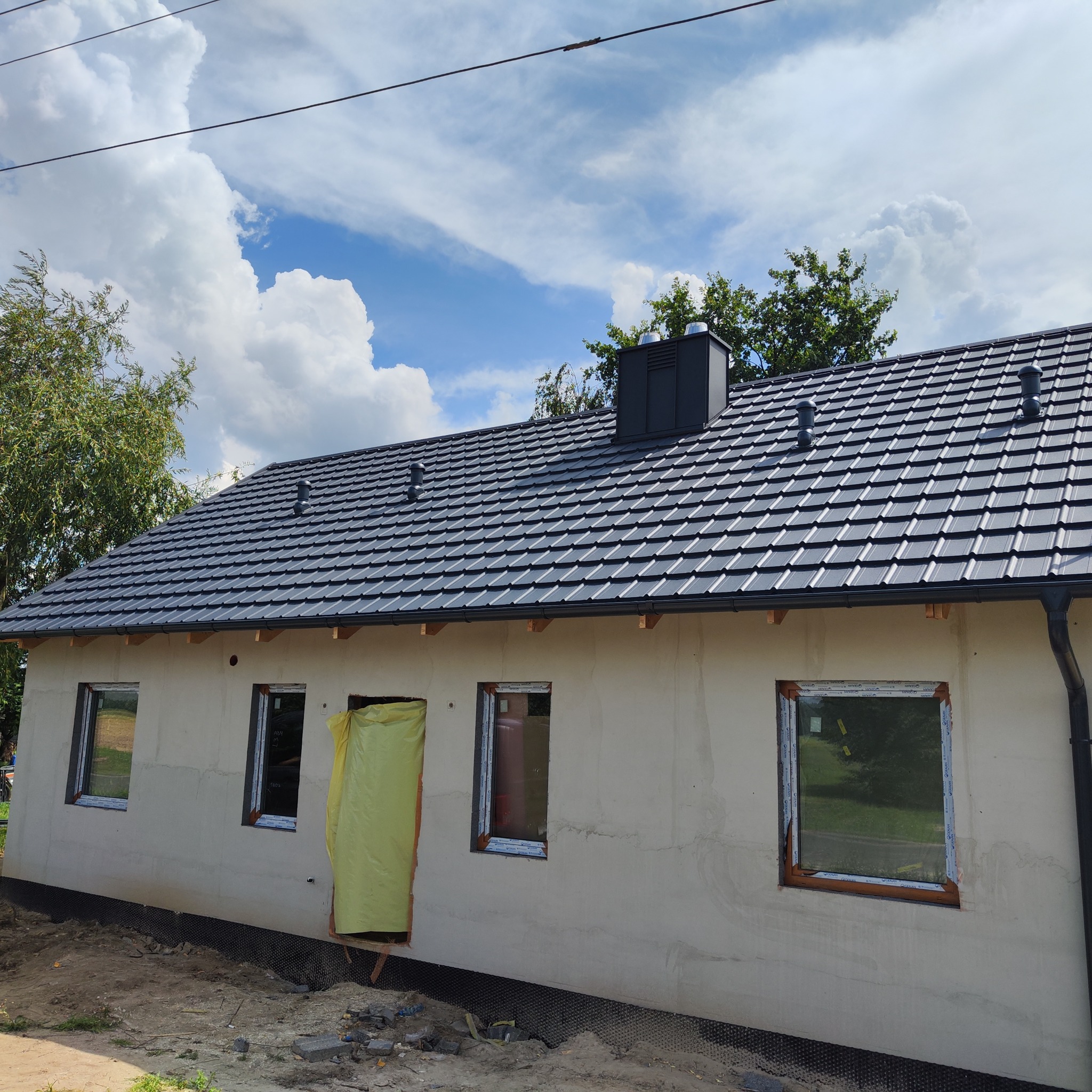Dom jednorodzinny w trakcie budowy: nowy, grafitowy dach z blachodachówki, okna wstawione, elewacja surowa, widoczne elementy wentylacji dachowej.