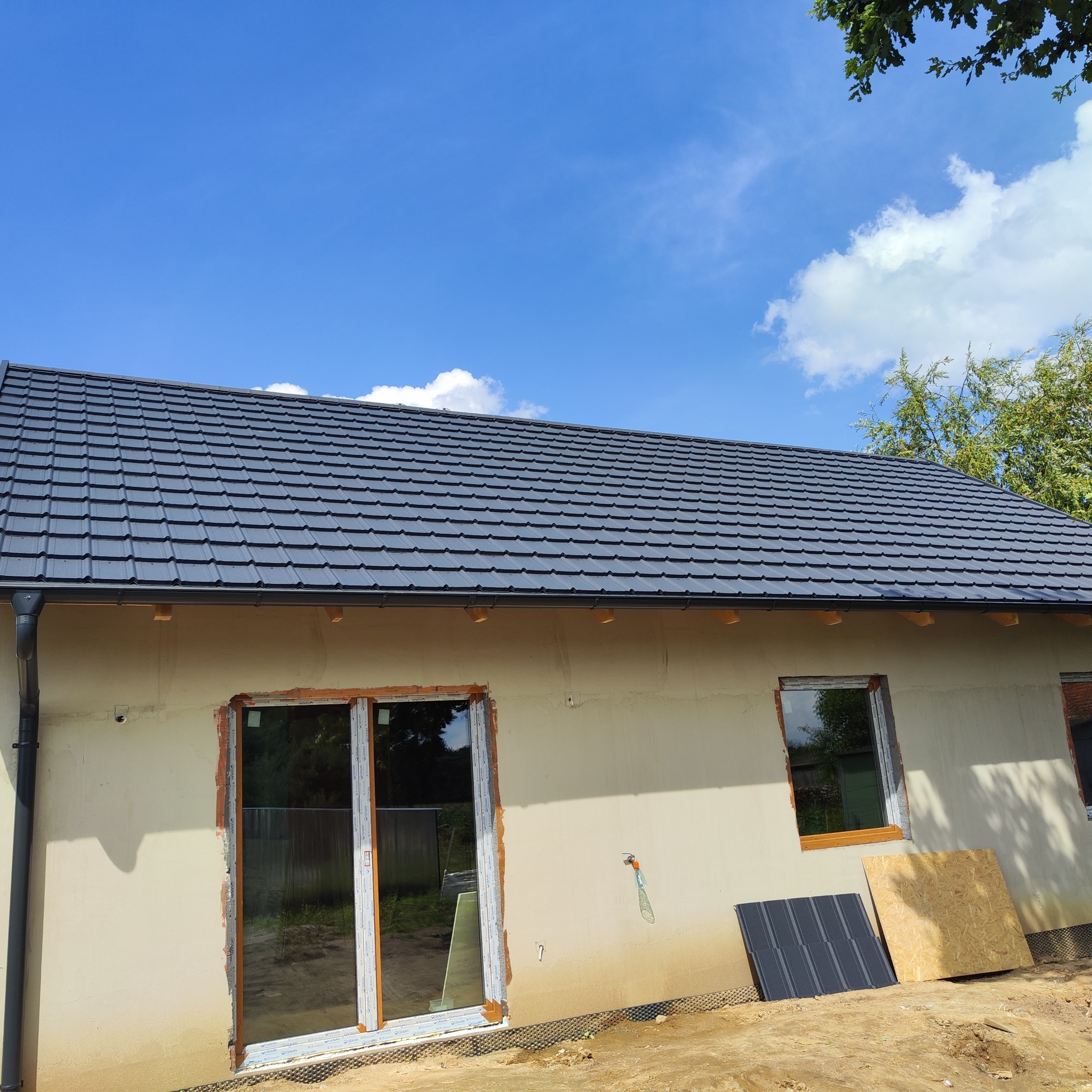 Dom w trakcie budowy z nowym, grafitowym dachem z blachodachówki. Widoczne okna i drzwi balkonowe. Na ziemi leżą płyty i arkusz blachy.