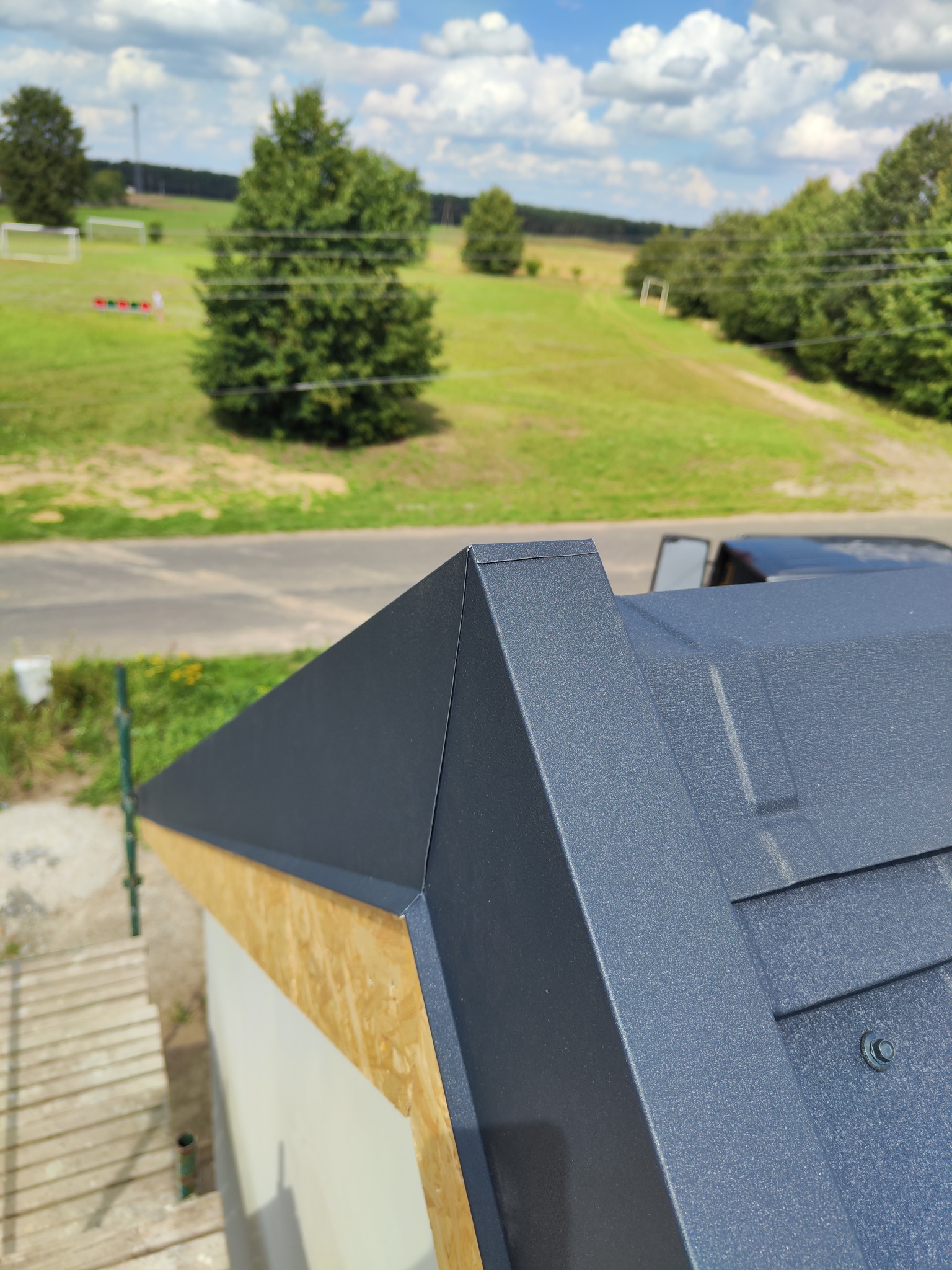 Szczegółowe ujęcie fragmentu nowo krytego dachu z blachy na budynku z widocznym deskowaniem i elementami mocującymi na tle zielonego krajobrazu.