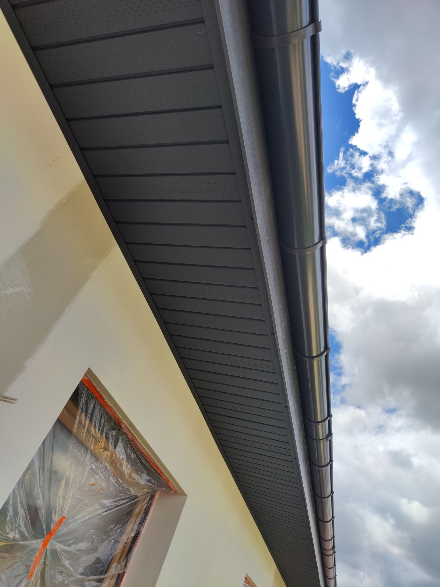 Szare panele dachowe z rynnami, widoczne z dołu na tle nieba z chmurami. Okno zabezpieczone folią malarską. Ukośne ujęcie.