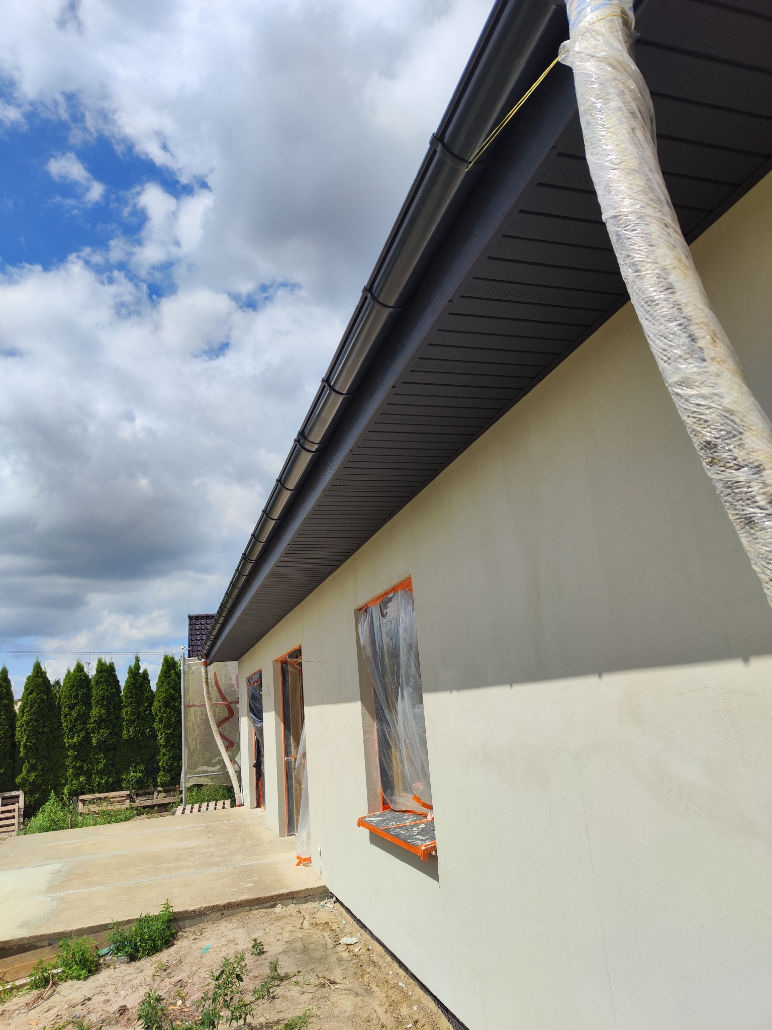 Budynek jednorodzinny w trakcie prac wykończeniowych elewacji, z widocznymi oknami zabezpieczonymi folią i elementami dachu. Tło: błękitne niebo.