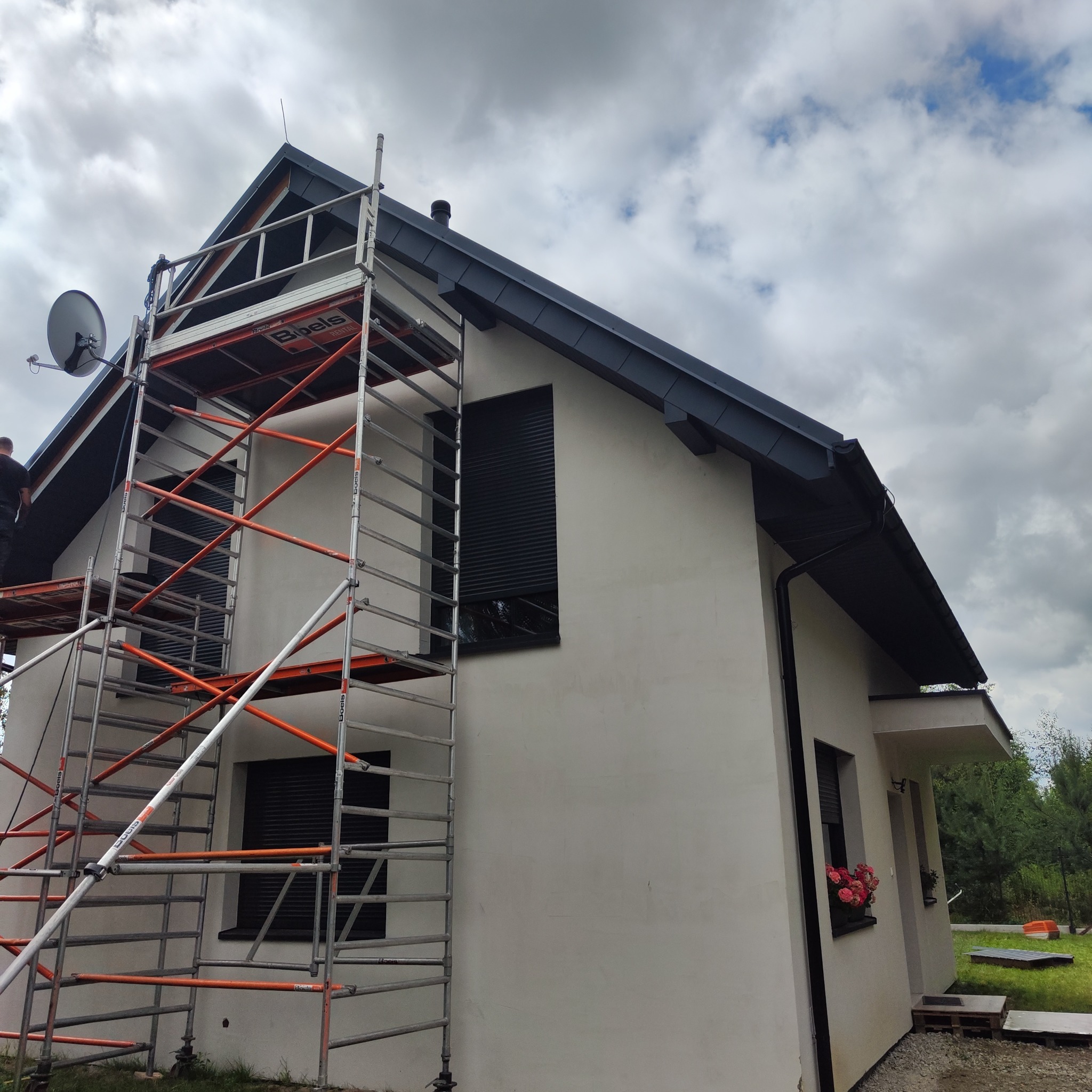 Dom jednorodzinny z nowym, grafitowym dachem z blachy na rąbek stojący, widoczny ze stojącego rusztowania na tle zachmurzonego nieba.