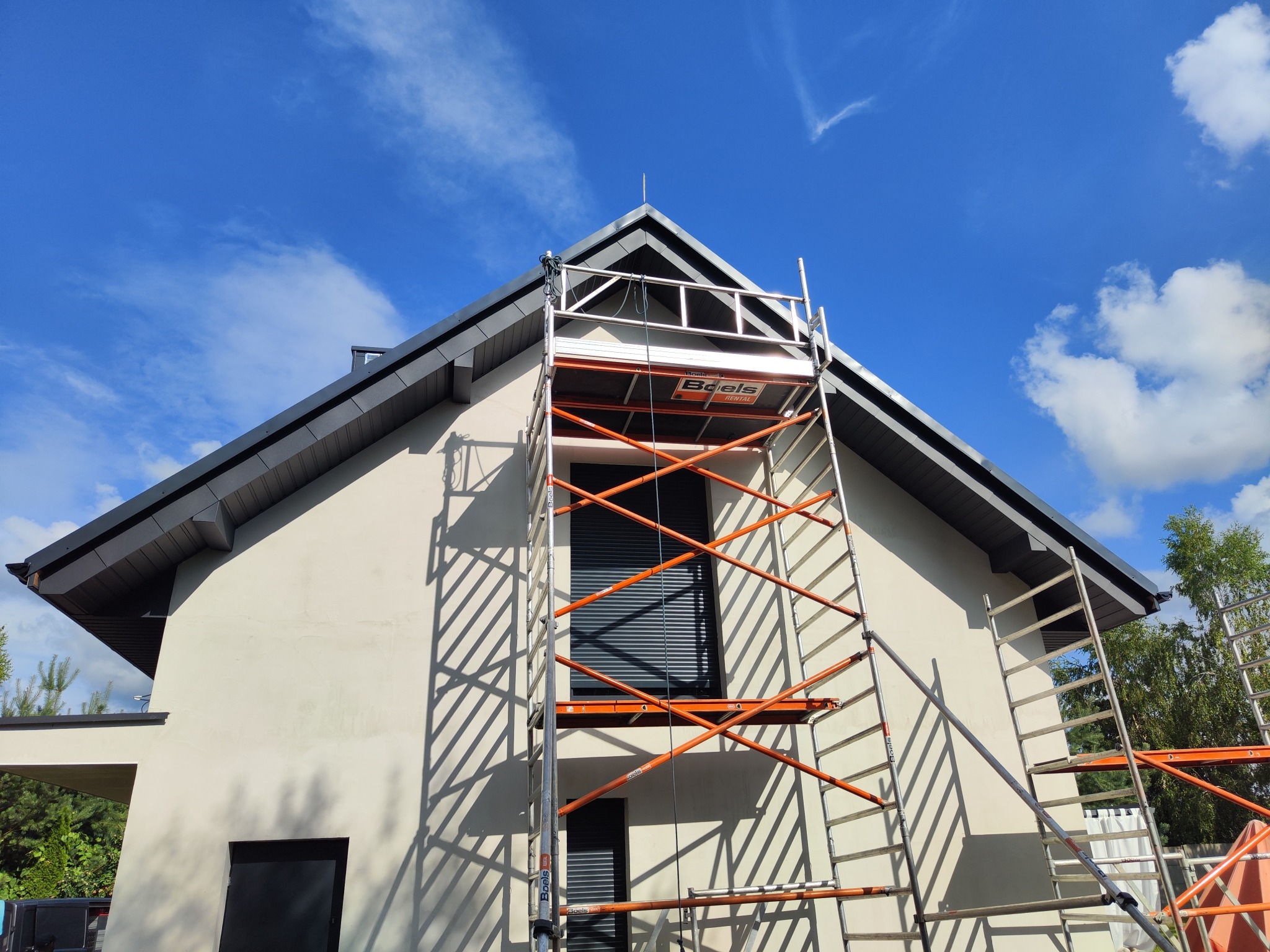 Dom jednorodzinny z ciemną dachówką, widziany od dołu. Na pierwszym planie rusztowanie z platformą roboczą, błękitne niebo z chmurami w tle.