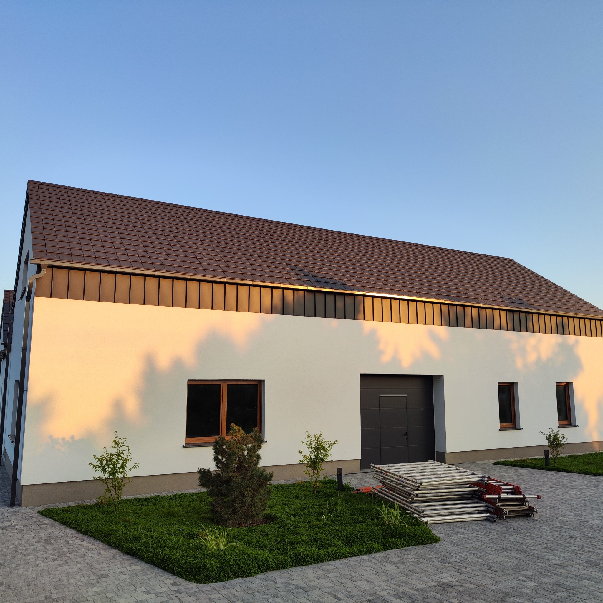 Nowoczesny dom z brązowym dachem i stosem rusztowań przy ścianie w promieniach słońca. Widoczna kostka brukowa i zieleń. Minimalistyczny design.