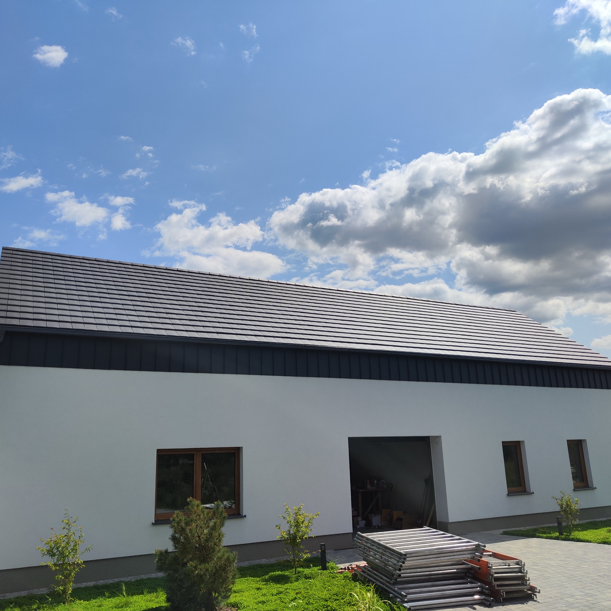 Nowoczesny dom z ciemnoszarą dachówką ceramiczną na jasnej elewacji. Widoczne okna z drewnianą stolarką, trawnik i rusztowania. Błękitne niebo z chmurami w tle.