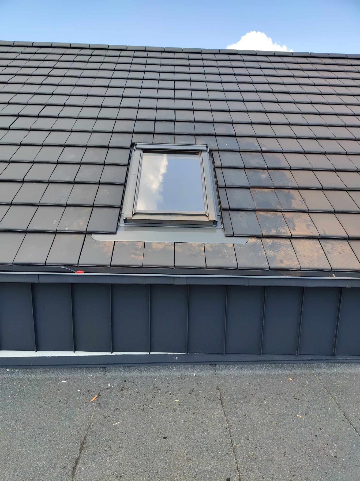 Czarny dach z dachówki ceramicznej z oknem dachowym, odbijającym błękit nieba i obłoki. Widoczny fragment orynnowania i pokrycia dachu.