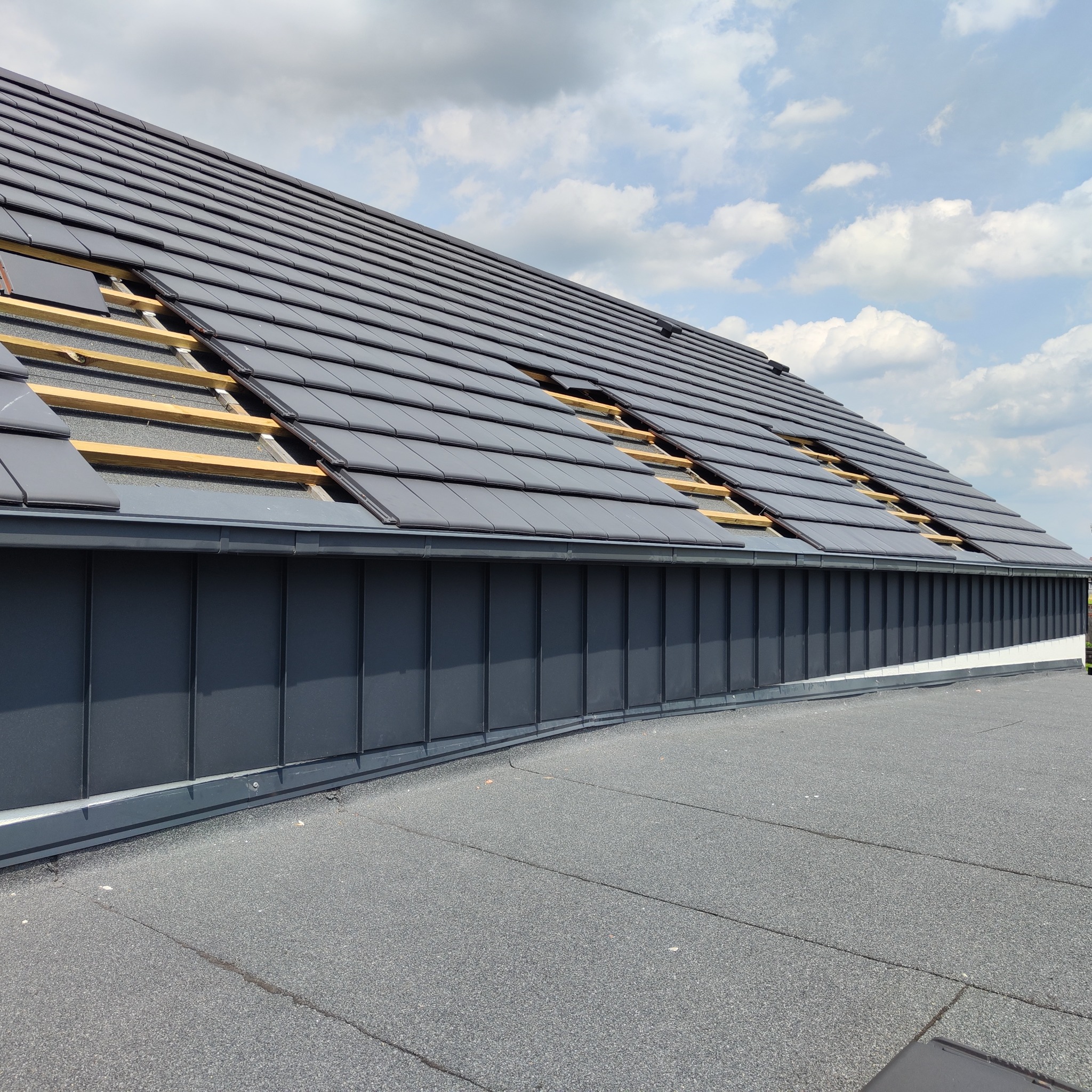 Częściowo pokryty dach z ciemnej dachówki ceramicznej, widoczne drewniane łaty, błękitne niebo z obłokami w tle. Fragment pokrycia papą na płaskim dachu.