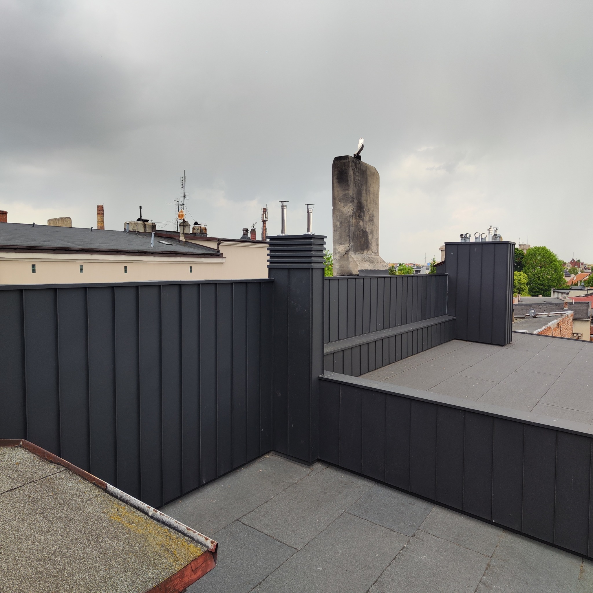Nowoczesne, czarne obicie pionowe dachu z blachy na tle miejskiego krajobrazu i pochmurnego nieba. Widoczne kominy i inne budynki.