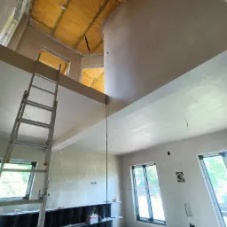 Wnętrze domu w trakcie prac wykończeniowych, widok na wysoki sufit z nałożonymi świeżymi tynkami, zabezpieczona folią przestrzeń pod dachem, aluminiowa drabina oparta o ścianę, widoczne okna...